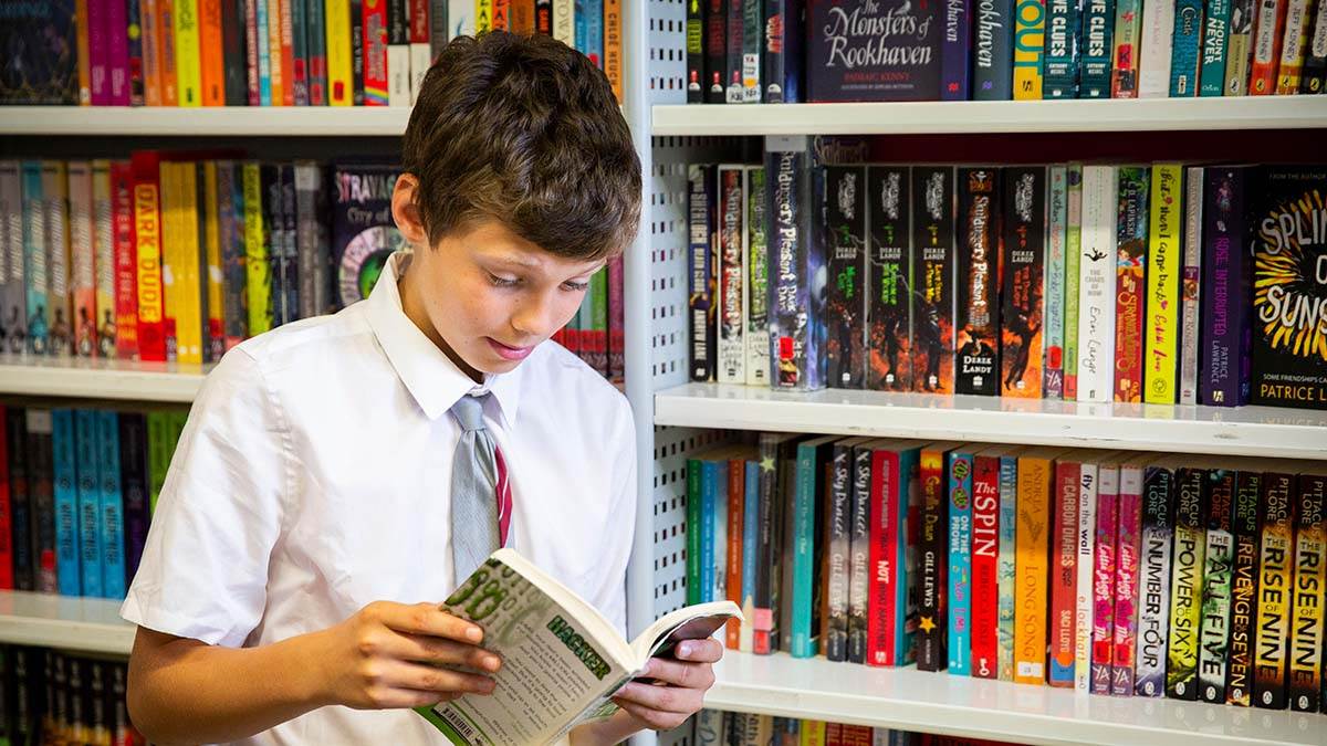 A photo of a boy reading by bookshelves in his school library