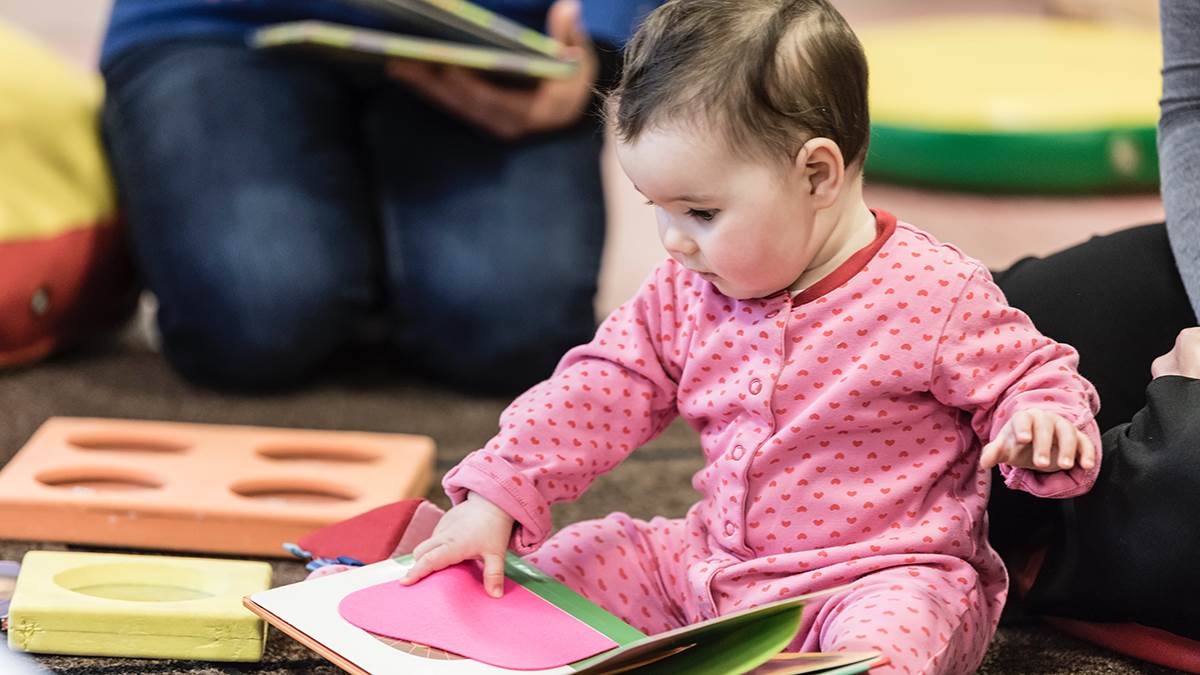 A photo of a young baby sitting on the floor of a children's centre playing with a book