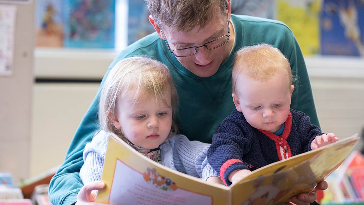 A photo of a man reading to a baby and a girl in a library