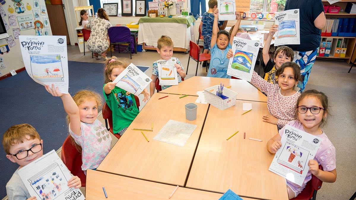 Children in a classroom wearing pyjamas and excitedly holding up Pyjamarama activity sheets