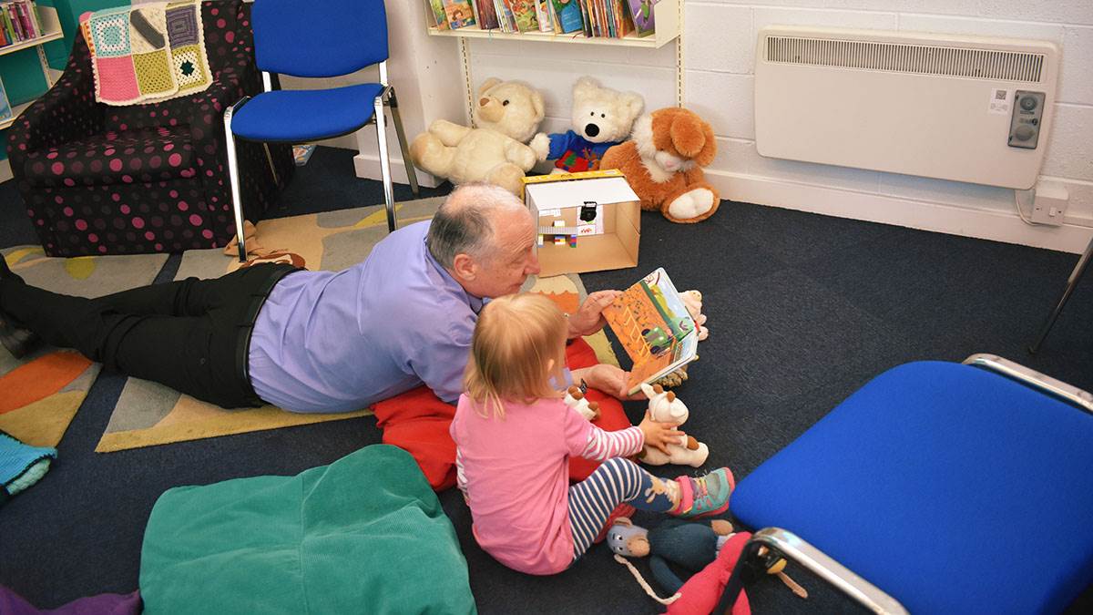 Librarian Paul lying on the floor of the library sharing an open book with a young child