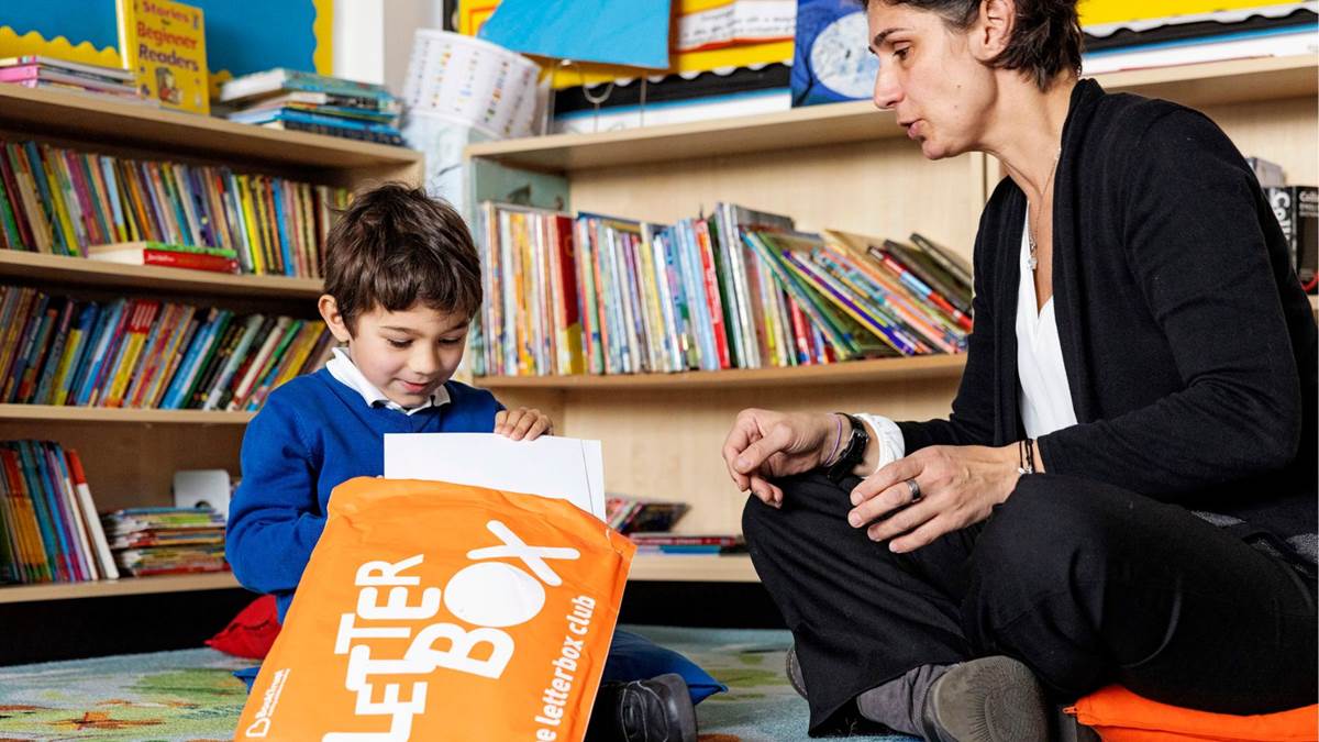 A teacher sitting on the floor of a school book corner with a child excitedly opening a Letterbox Club parcel