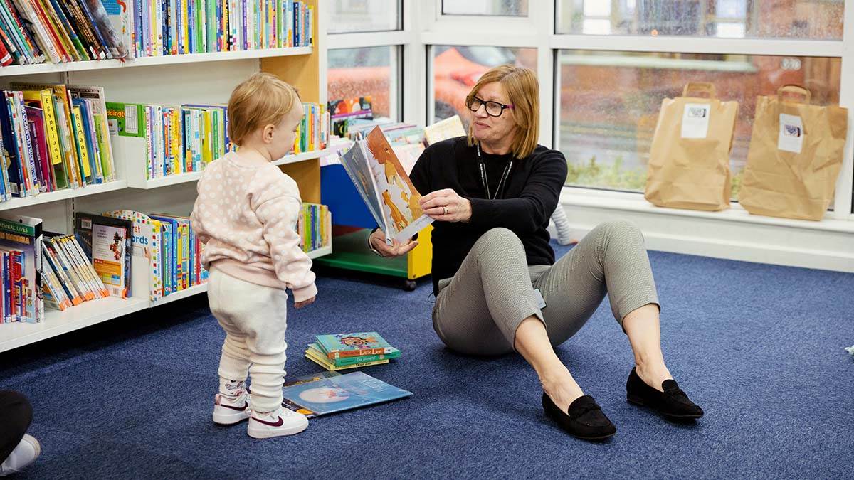 A young child wandering over to a librarian reading a story, looking interested
